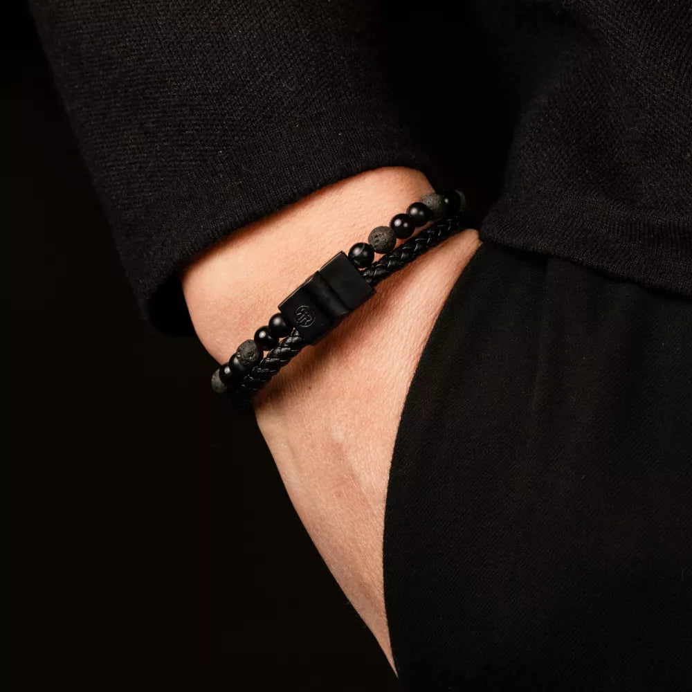 Een persoon in een zwarte trui en broek heeft zijn hand in zijn zak en laat de Rebel & Rose Full Metal Combo 12mm Black Rocks heren kralen armband met kralen en gevlochten details om zijn pols zien.