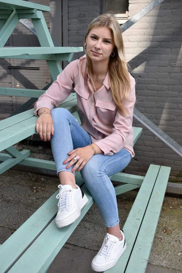 Een vrouw met lang blond haar ontspant op een mintgroene bank, haar roze blouse en lichtblauwe jeans matchen met haar witte sneakers. Haar Ring Twee ronde vormen Goud|Zand R5G van CLIC Creations glinstert terwijl een nabijgelegen metalen structuur het unieke, handgemaakte ontwerp uit Nederland verlicht.