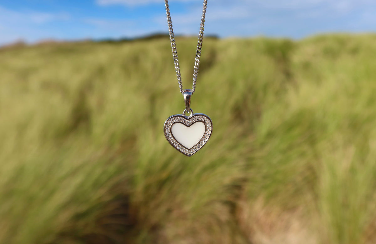 De Demi | AsHanger massief | Zirkonia van Juwelier van der Kooij heeft een zilveren hanger in de vorm van een hart met zirkonia's en een wit hart. De hanger staat scherp af tegen het hoge groene gras en een blauwe lucht.
