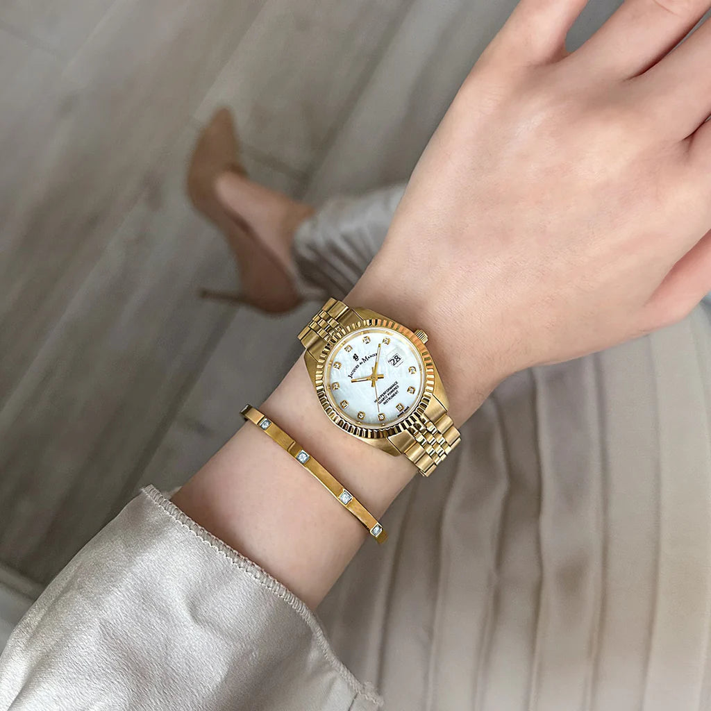 Een persoon die de Jacques Du Manoir Inspiration 31mm met witte wijzerplaat en gouden armband draagt, paart deze met lichtgekleurde kleding en beige hoge hakken terwijl ze op een lichte houten vloer staat.