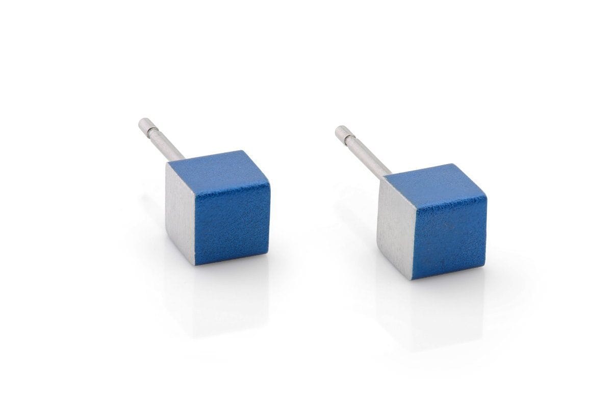 De Oorsieraad Kubusjes Blauw O28B van CLIC Creations bestaat uit twee kubusvormige oorbellen met blauwe en zilveren vlakken, elk op een klein staafje. Deze oorbellen zijn handgemaakt in Nederland en hebben een uniek ontwerp, omdat ze rusten op een wit oppervlak en licht reflecteren.