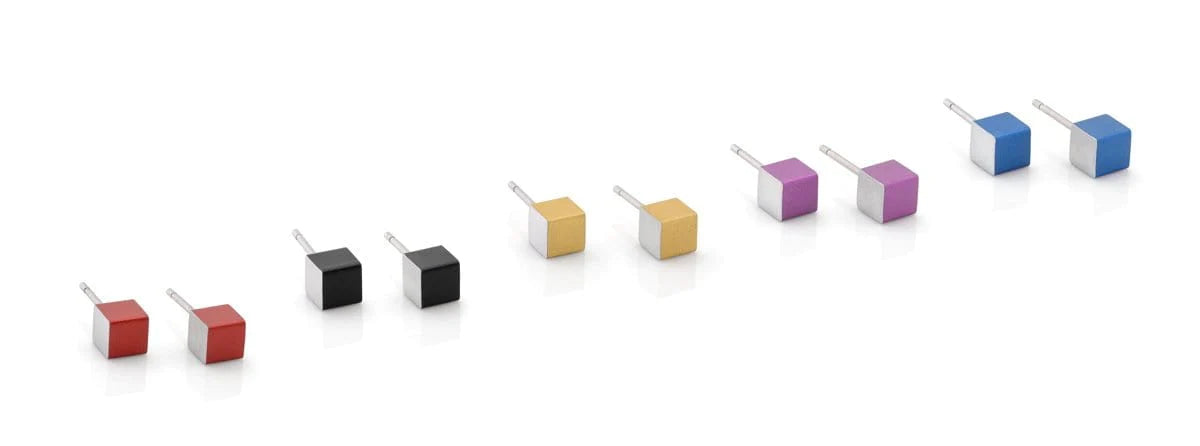 Een rij CLIC Creations USB-laders met een uniek kubusvormig ontwerp in levendige kleuren: twee rode, twee zwarte, één gele, één gouden, één paarse (Oorsieraad Kubusjes Paars O28P) en twee blauwe. Handgemaakt in Nederland op een reflecterend oppervlak met elke USB-pen uitgeschoven.