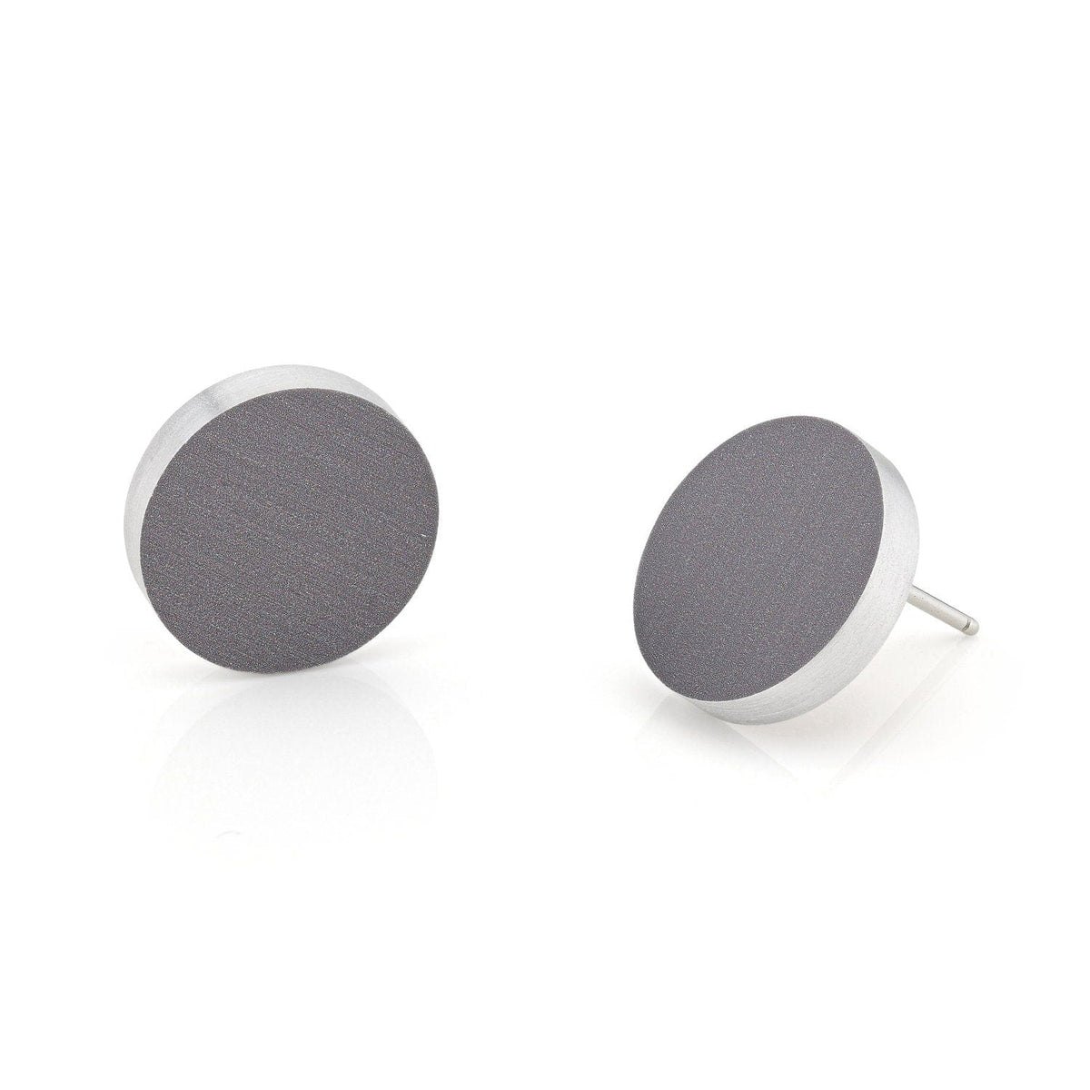 De Oorsteker rond O53AG oorbellen van CLIC Creations zijn handgemaakt in Nederland en hebben een glad donkergrijs oppervlak met zilveren randen. Eén oorbel staat rechtop, terwijl de andere gekanteld is om de pin te onthullen tegen een schone, reflecterende witte achtergrond.