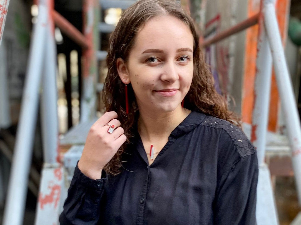 Een vrouw met krullend haar in een zwarte blouse lacht voor metalen leuningen, met de CLIC Creations Oorhanger lange staafjes Rood O38R en een ring om, met één hand bij haar nek. De stedelijke achtergrond is voorzien van licht wazige, rustieke steigers.
