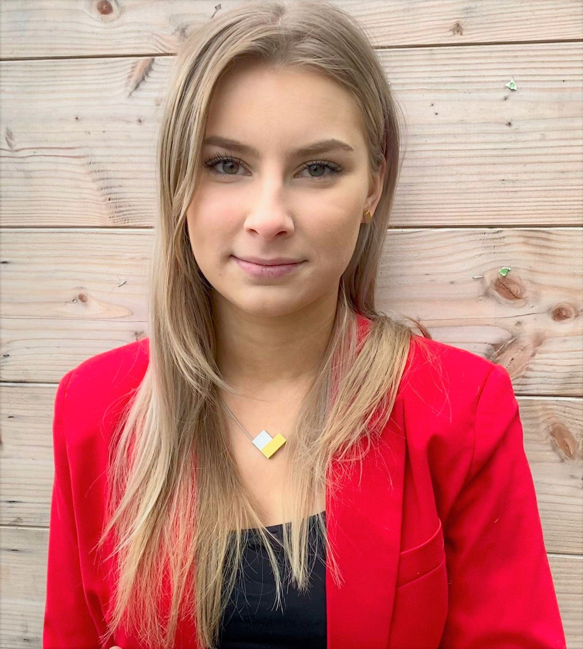 Een persoon met lang blond haar draagt een felrode blazer over een zwart topje en de CLIC Creations Collier V-vorm Rood C150R ketting, die een uniek ontwerp en magnetische sluiting heeft. Ze staan tegen een lichte houten muur en kijken recht in de camera.