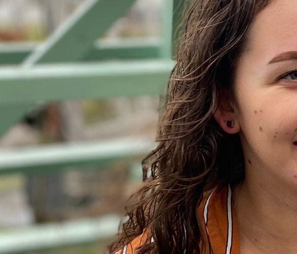 Een close-up van een vrouw met golvend bruin haar en een oranje en wit gestreepte kraag toont de Oorsieraad Kubusjes Zwart O28Z oorbel van CLIC Creations, handgemaakt in Nederland. Ze heeft zichtbare sproeten en de wazige achtergrond bevat groene metalen structuren. Slechts de helft van haar gezicht is zichtbaar.