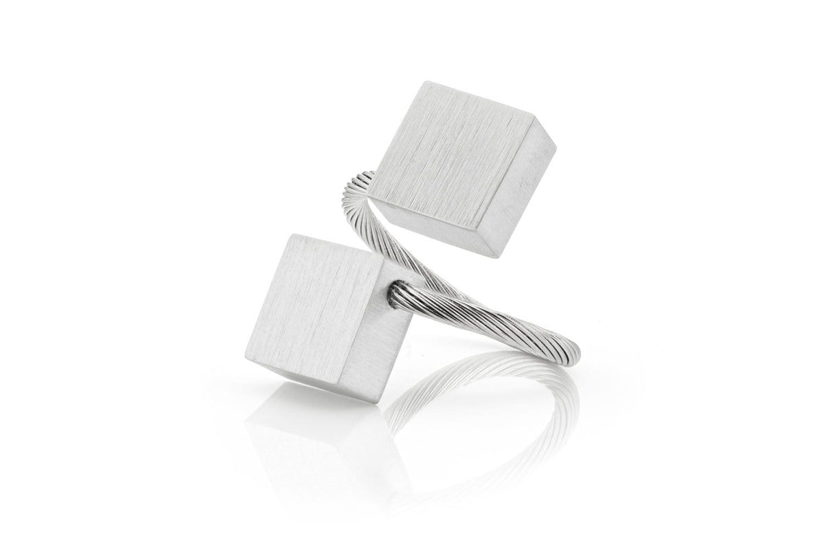 De Ring Vierkanten R6 van CLIC Creations is een minimalistisch metalen sculptuur, handgemaakt in Nederland. Het bestaat uit twee kubusvormige elementen die met elkaar verbonden zijn door gedraaide draad en heeft een zilveren afwerking, elegant gebalanceerd op een reflecterend oppervlak, wat lijkt op een perfect vervaardigde ring.