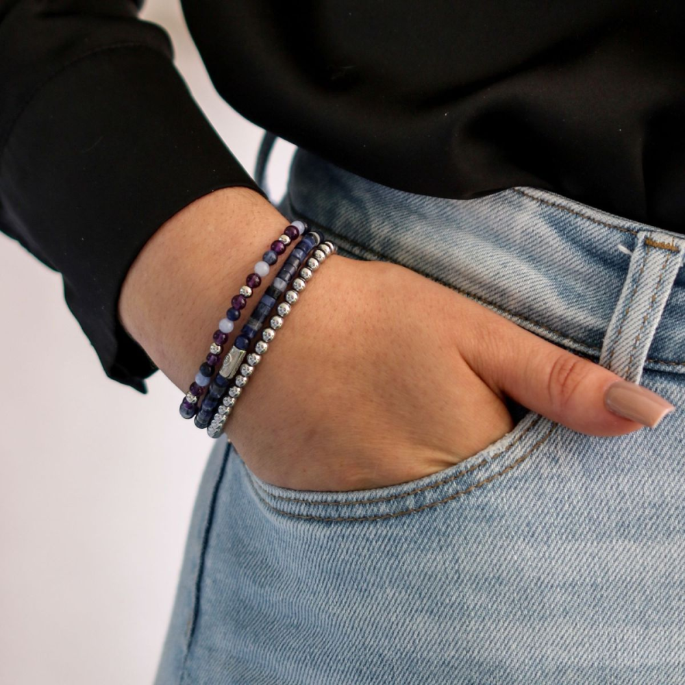 Een persoon in een lichtblauwe spijkerbroek en een zwart overhemd heeft zijn hand in zijn zak en onthult drie kralenarmbanden - een daarvan is de Rebel & Rose Silver Shine - 4mm Kralen Armband Dames - in blauwe en zilveren tinten om zijn pols.