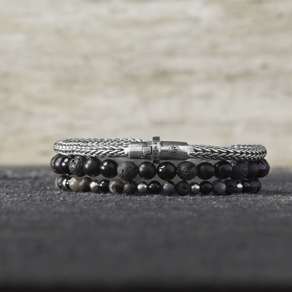 De Shiny Night - 4mm Kralen Armband Dames van Rebel & Rose wordt gestapeld getoond, met zwarte edelsteen en zilveren kralen plus een geweven zilveren ketting met sluiting, weergegeven op een donker gestructureerd oppervlak tegen een vage beige achtergrond.