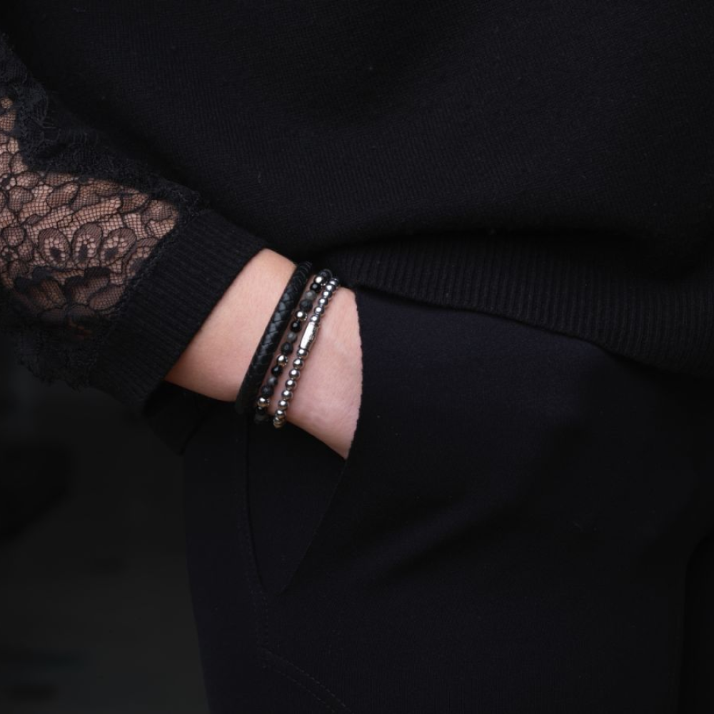 Een persoon in een zwarte outfit met kanten mouwen heeft één hand in zijn zak en toont een Rebel & Rose Shiny Night - 4mm - kralen armband dames om zijn pols.