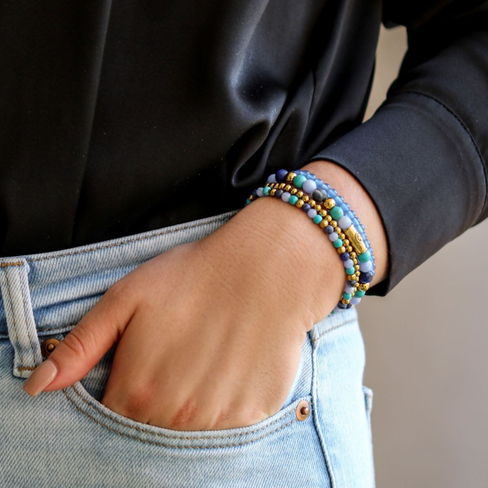 Een persoon met zijn hand in zijn zak pronkt met een Rebel & Rose Winter Blues 4 mm armband, met turquoise en blauwe agaatkralen met een 18 kt geelgouden ion-plating, in combinatie met een zwart shirt met lange mouwen en een lichtblauwe jeans.