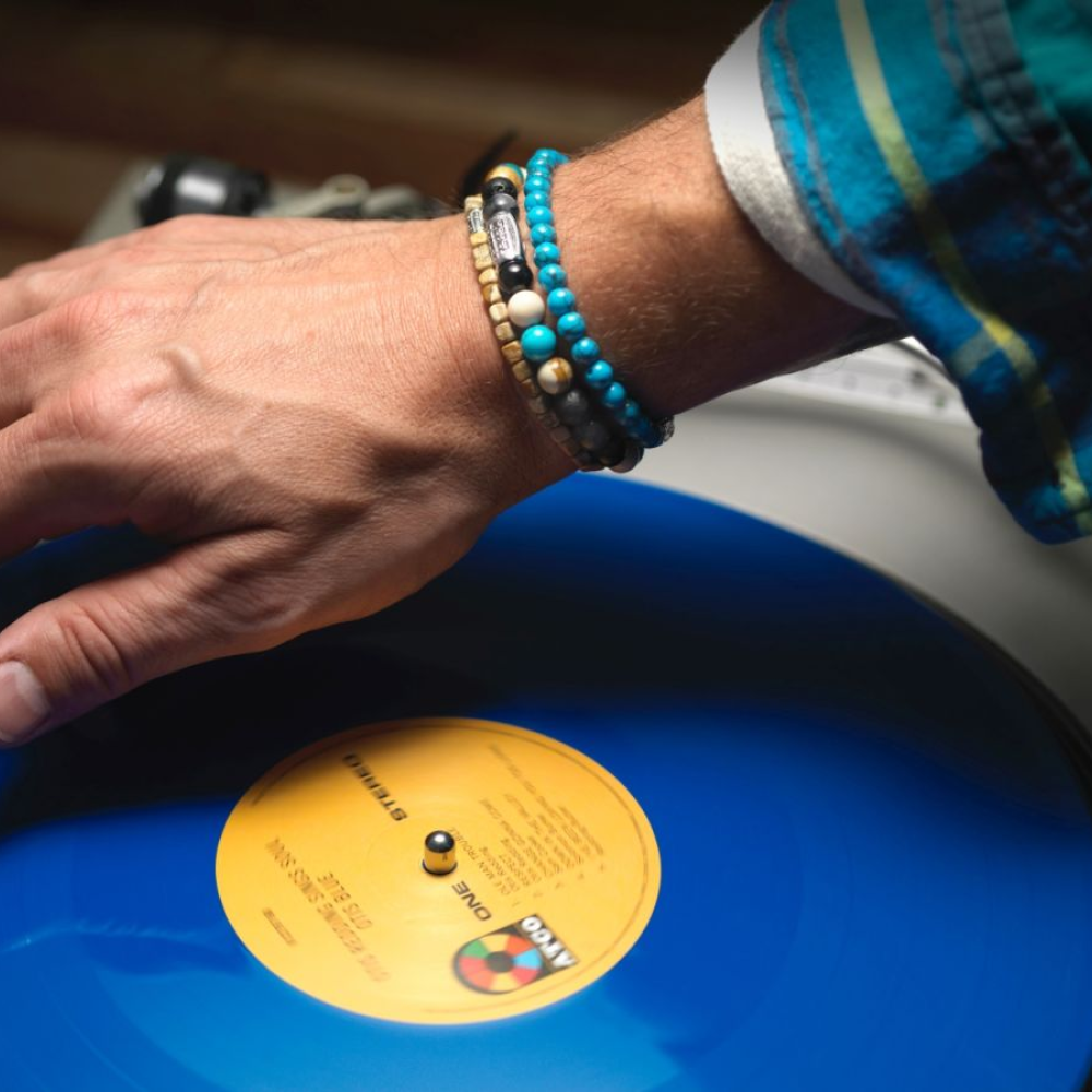 Een persoon met kleurrijke kralenarmbanden, waaronder de Rebel & Rose Roll The Dice - Woodstock - 4mm Heren Kralen Armband, en een blauwgroen shirt draait een blauwe vinylplaat met een geel label op een platenspeler.