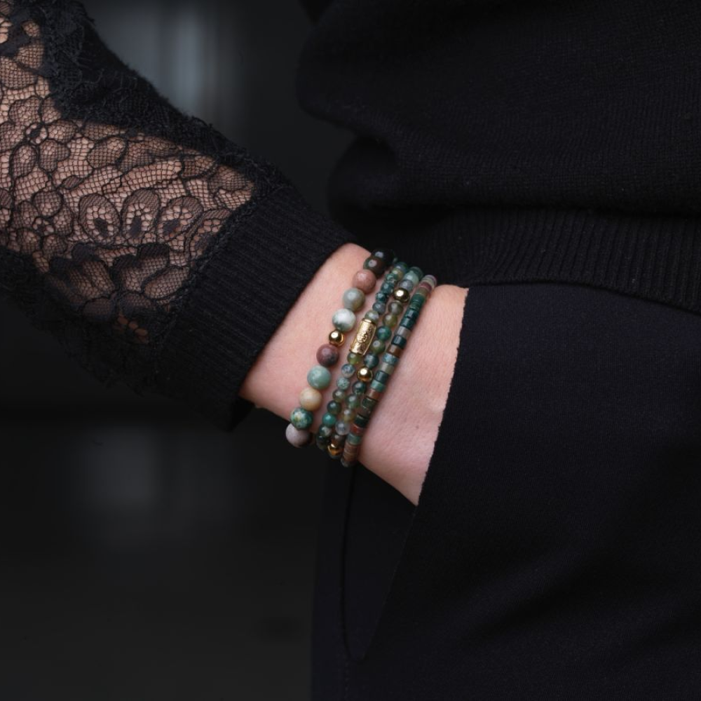Een persoon met één hand in zijn zak draagt een donkere broek en een top met zwarte kanten mouwen. Om zijn pols draagt hij kralenarmbanden van Rebel & Rose, waaronder de Spring Shine 18 kt geelgouden geïoniseerde 6 mm Kralen Armband Dames.