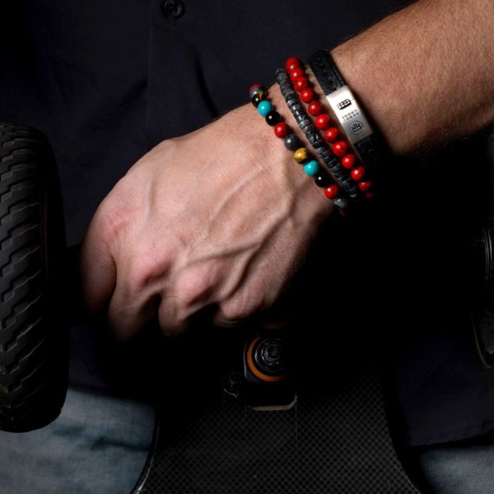 Een close-up van een hand die een skateboard vasthoudt, gestyled met kleurrijke kralenarmbanden en de Rebel & Rose Slices - Grey Seduction - 6mm Heren Kralen Armband. De donkere achtergrond contrasteert met het donkere shirt en de spijkerbroek van de drager.