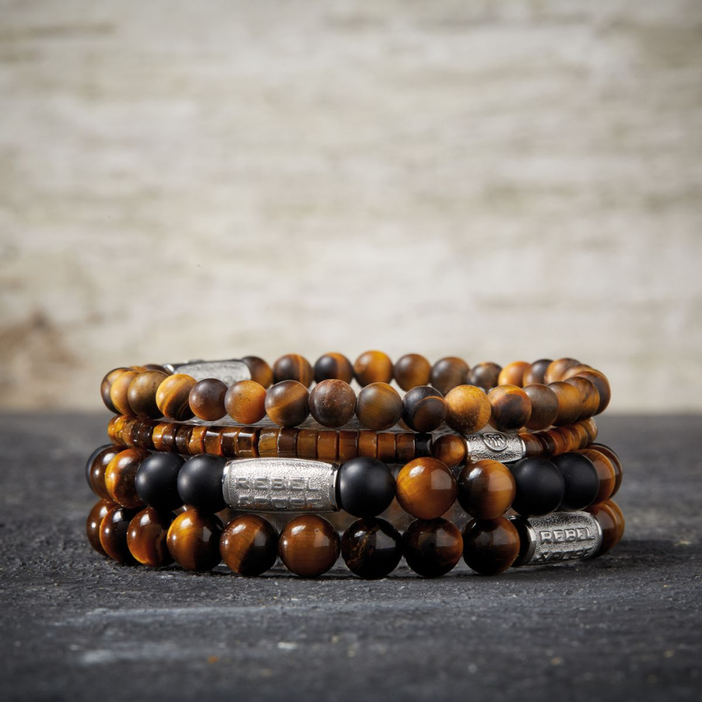Drie kralenarmbanden, waaronder de Rebel & Rose Tiger Lily - 8mm Kralen Armband Heren in bruin, zwart en goud met zilveren accenten, zijn gestapeld op een donker stenen oppervlak - perfect als herenarmband.