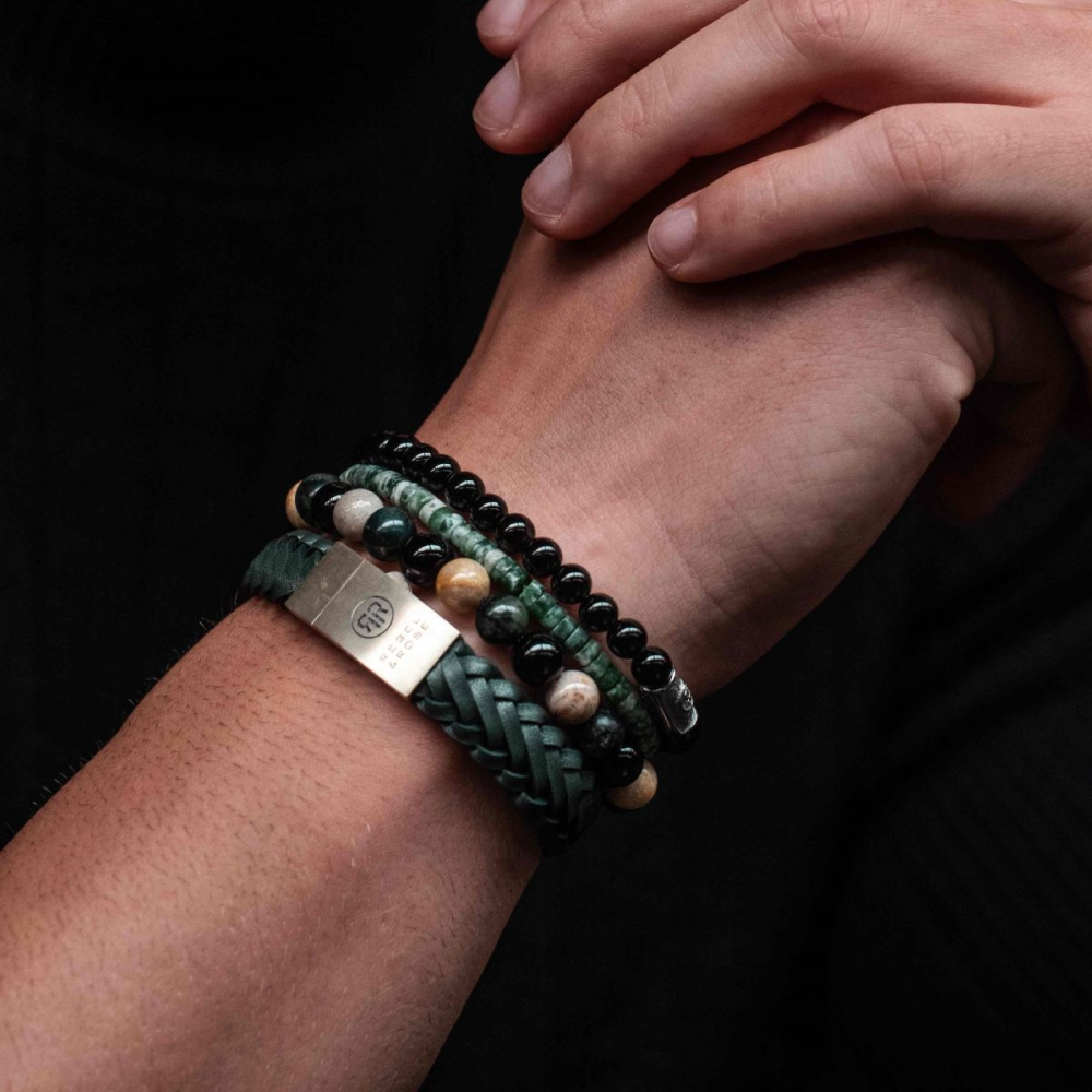 De handen van een persoon worden getoond, de ene houdt zachtjes de andere pols vast, versierd met de Rebel & Rose Autumn Storm - 8mm Kralen Heren Armband, met kralen in groene, zwarte en taankleurige tinten.