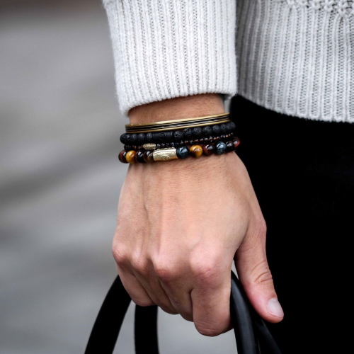 Een persoon in een lichtgrijze trui houdt een zwart handvat vast en toont gestapelde armbanden waaronder de Rebel & Rose Black Moon - All-Black 8mm Heren Kralen Armband om zijn pols tegen een wazige achtergrond.