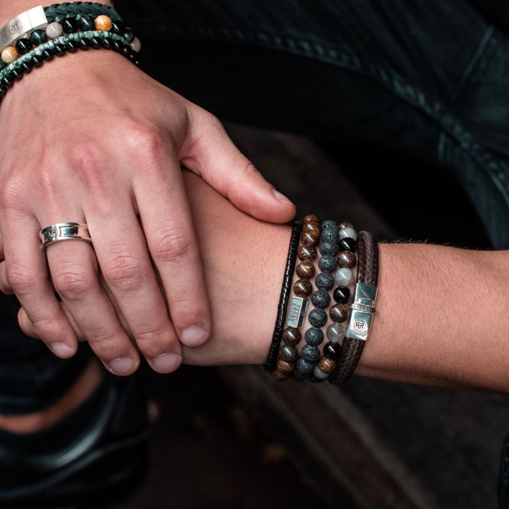 Een close-up van twee handen, de ene met een zilveren ring en een Rebel & Rose Black Moon - All-Black - 8mm Heren Kralen Armband, de andere met gelaagde kralen, leer en metalen armbanden, beide rustend op een knie.