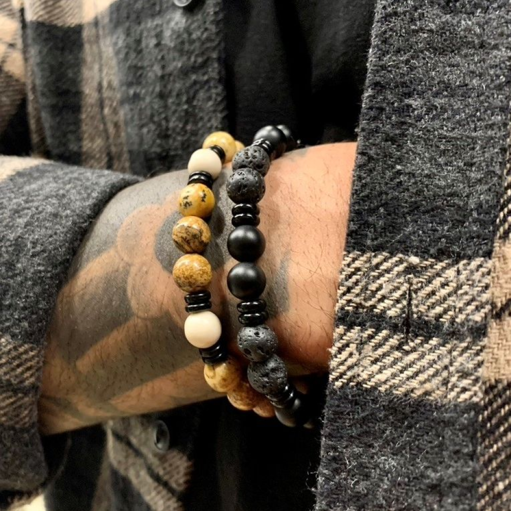 Een getatoeëerde persoon draagt twee kralenarmbanden - de ene is de Rebel & Rose Woodstock 8mm Kralen Heren Armband en de andere heeft bruine en crèmekleurige kralen - in combinatie met een grijs en beige geruit overhemd of jasje.