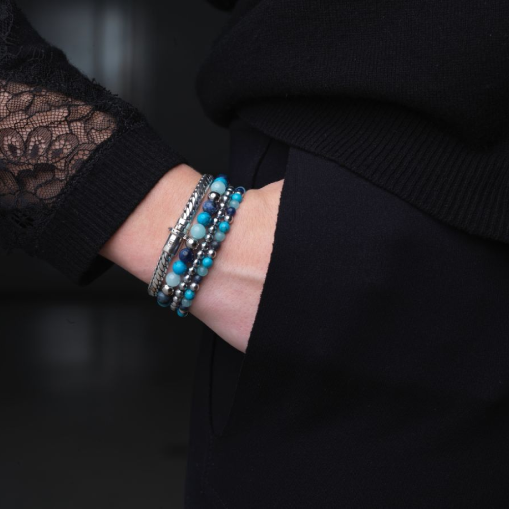 Een persoon in een top met zwarte kanten mouwen showt gestapelde blauwe, turquoise en zilveren kralenarmbanden met de stijlvolle Rebel & Rose Apollo Small six - 18,30 gr. 925 zilveren armband; de donkere achtergrond is onscherp.