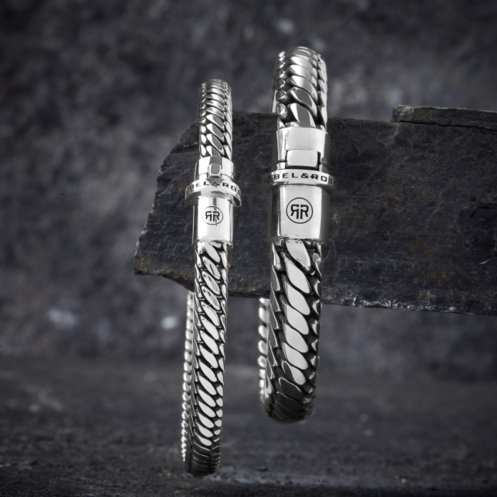 Twee zilveren schakelarmbanden van Rebel & Rose, waaronder de Apollo Small six (18,30 gr., 925 zilver), worden getoond op een getextureerde donkere steen - de ene hangt terwijl de andere rechtop staat.