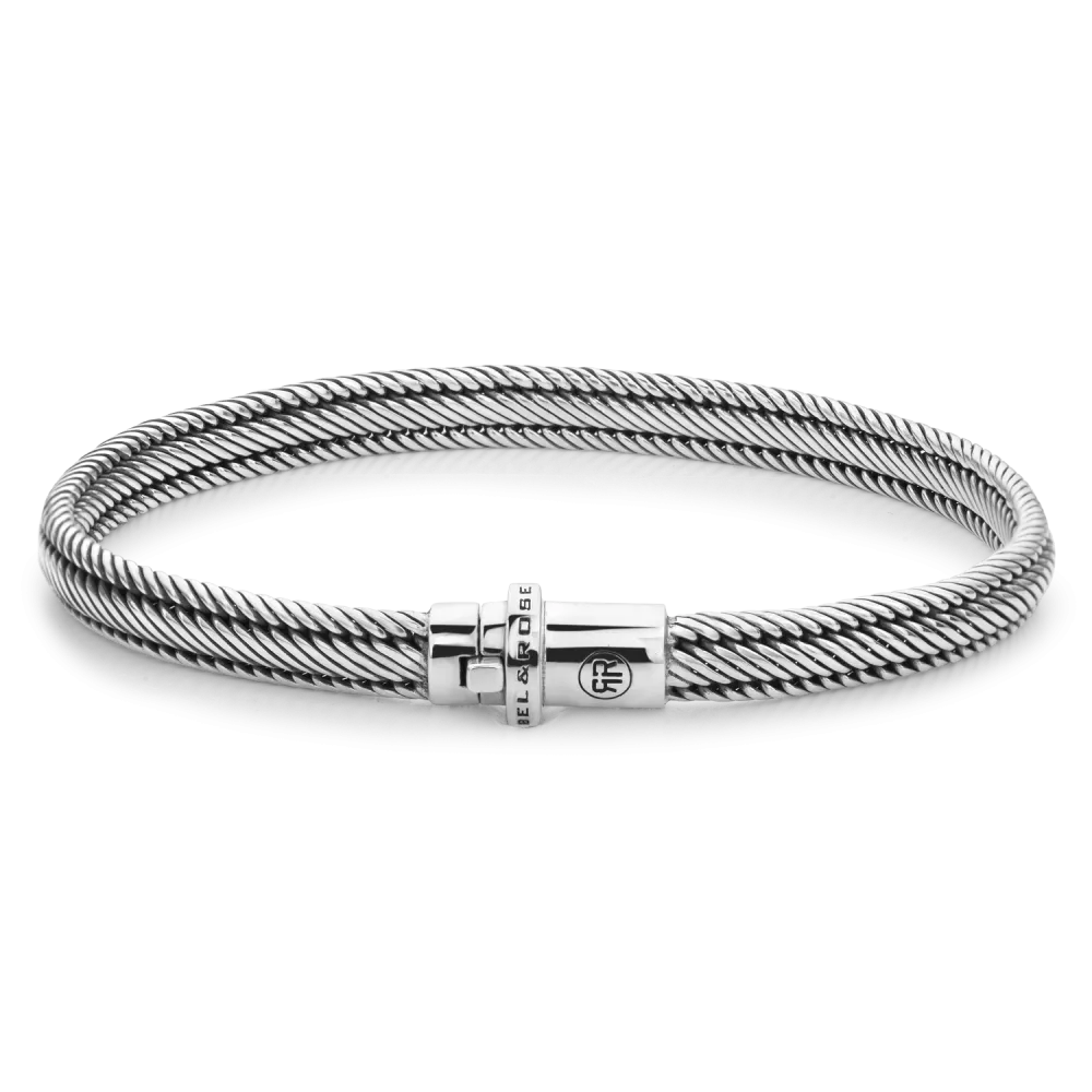 De Demeter Small Six armband van Rebel & Rose is een slank, zilverkleurig gedraaid kabelontwerp gemaakt van 17,11 gram 925 zilver, met een cilindrische sluiting gegraveerd met de merknaam en een cirkelvormig logo tegen een zwarte achtergrond.