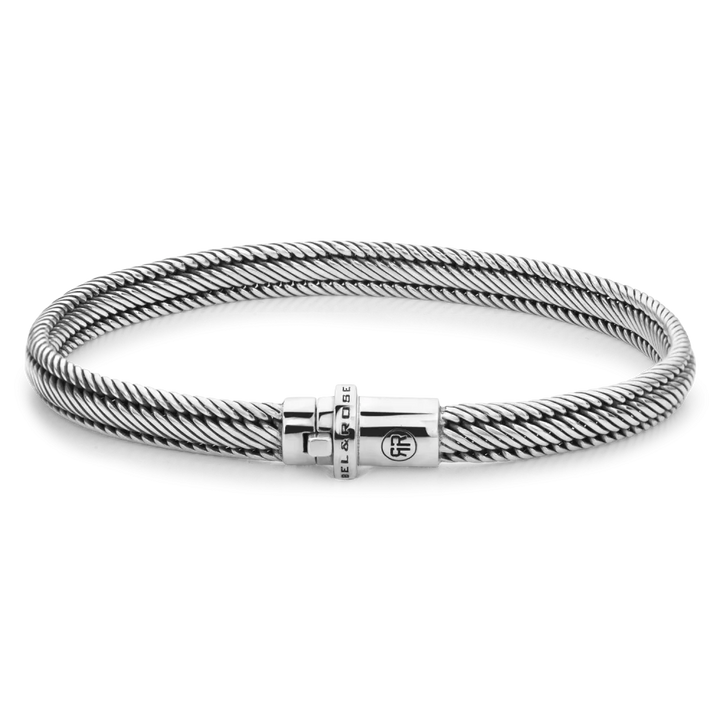 De Demeter Small Six armband van Rebel & Rose is een slank, zilverkleurig gedraaid kabelontwerp gemaakt van 17,11 gram 925 zilver, met een cilindrische sluiting gegraveerd met de merknaam en een cirkelvormig logo tegen een zwarte achtergrond.