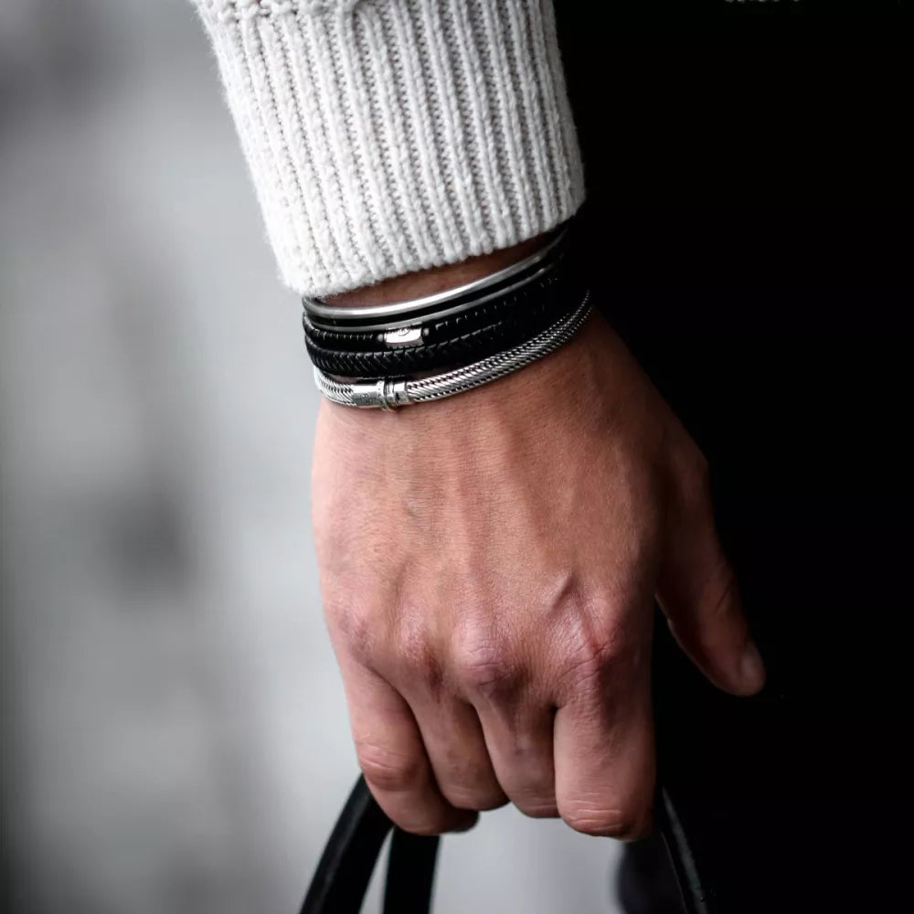 Een close-up van een hand die een zwart bandje vasthoudt, een grijze gebreide trui draagt en meerdere armbanden, waaronder de Rebel & Rose Demeter Small six - 17,11 gr. 925 zilver heren armband. De achtergrond is onscherp.