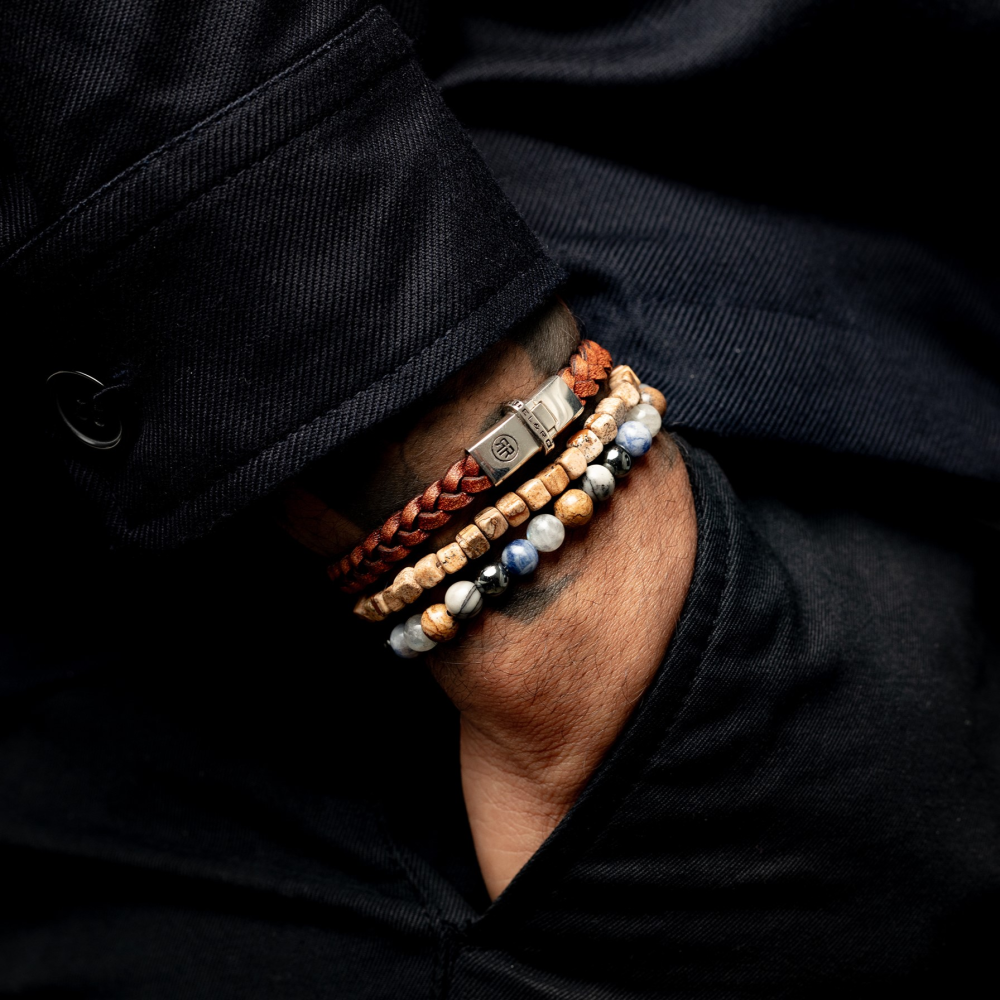 Een persoon draagt een donkere jas en vier armbanden, waaronder de Rebel & Rose Roll The Dice - Woodstock 4mm heren kralen armband, om zijn pols. Hun hand rust op hun been.