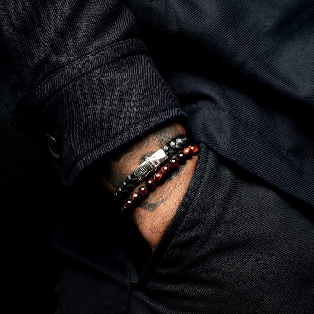 Een persoon met donkere lange mouwen en broek steekt zijn hand in zijn zak en laat twee kralenarmbanden zien - een zwarte en de Rebel & Rose Tiger Red Vintage - 8mm - Heren Kralen Armband om zijn pols.