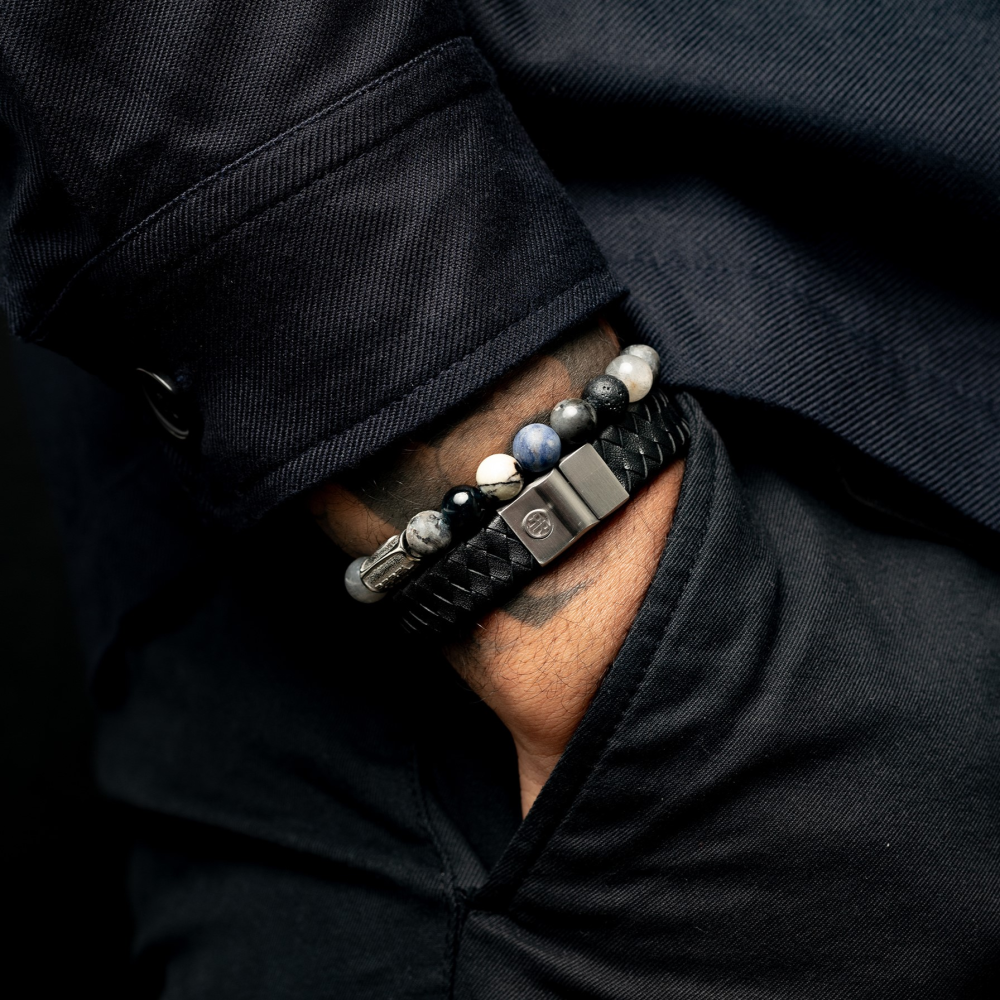 Een close-up van de onderarm van een persoon in een donkere jas, de hand in de zak, met twee stijlvolle Rebel & Rose armbanden - een kralen armband van Mountain High - 8mm Heren Kralen en een gevlochten - beide met slanke metalen sluitingen.