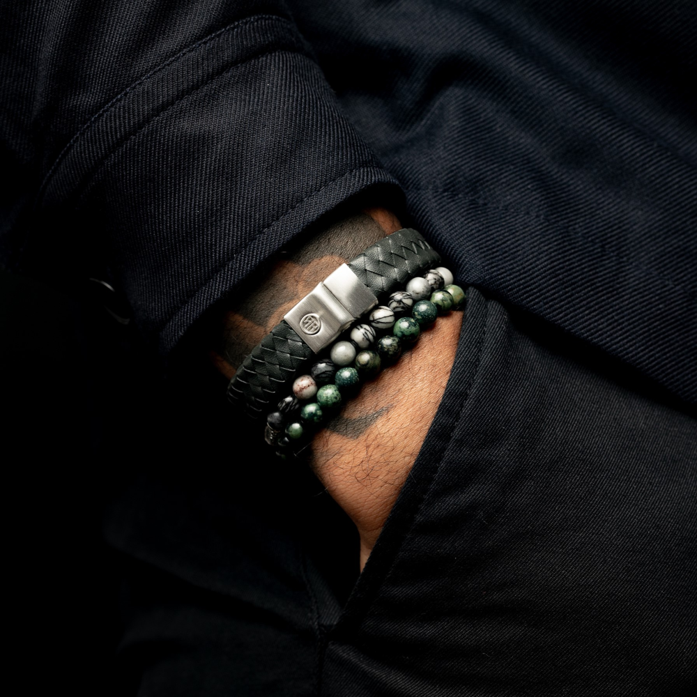 Een persoon in een donkere jas heeft zijn hand in zijn zak en laat de Rebel & Rose Black Wolf - 6mm Heren Kralen Armband zien: een zwart gevlochten leren armband gecombineerd met kralenarmbanden, waaronder een opvallend Black & Grey Agate ontwerp.