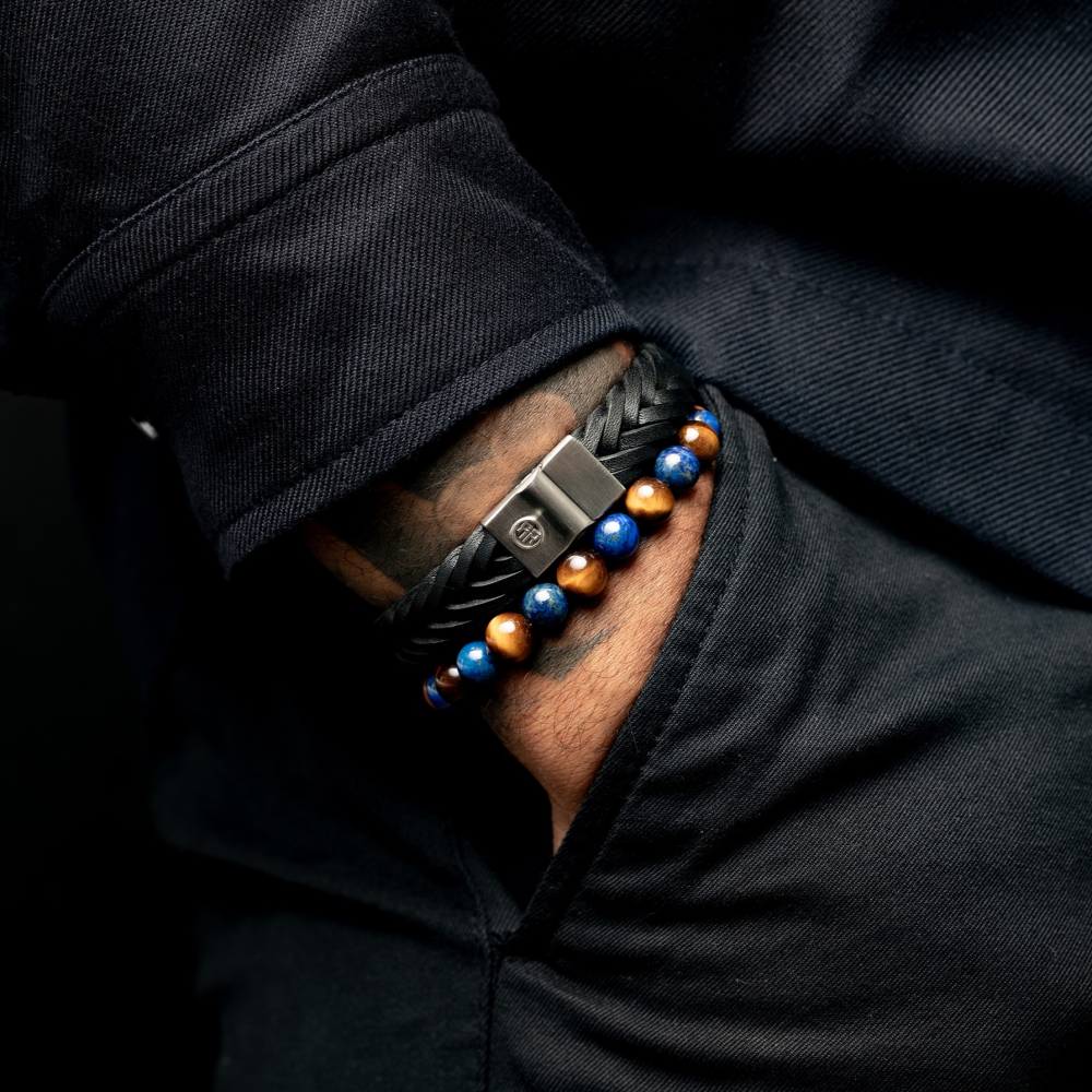 Een close-up van een pols met een luxe zwart gevlochten leren armband en de Rebel & Rose Mix Tiger Lapis - 8mm - Heren Kralen Armband, met een hand nonchalant weggestopt in een donkere zak.