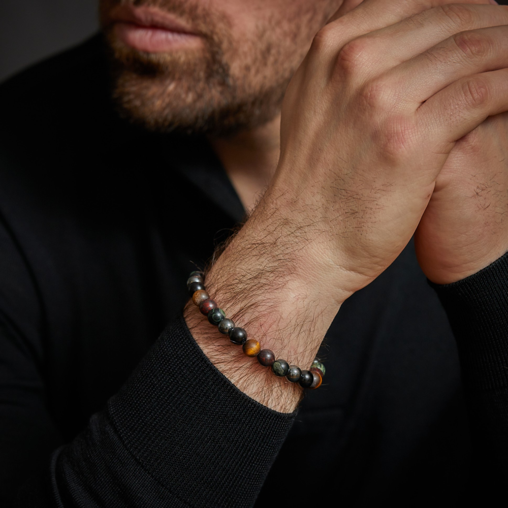 Een bebaarde man in een zwart overhemd houdt zijn handen tegen elkaar en laat op elegante wijze een Rebel & Rose Stormy Weather Vintage - 8mm heren kralen armband om zijn pols zien.