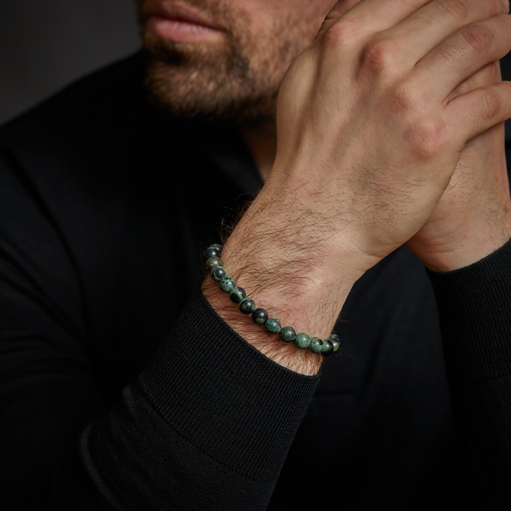 Een moderne man in een zwart shirt met lange mouwen laat zijn handen rusten en benadrukt de Rebel & Rose Green Rocks - 8mm Heren Kralen Armband om zijn pols. Zijn netjes getrimde baard en gedeeltelijk zichtbare gezicht benadrukken zijn stijlvolle look.