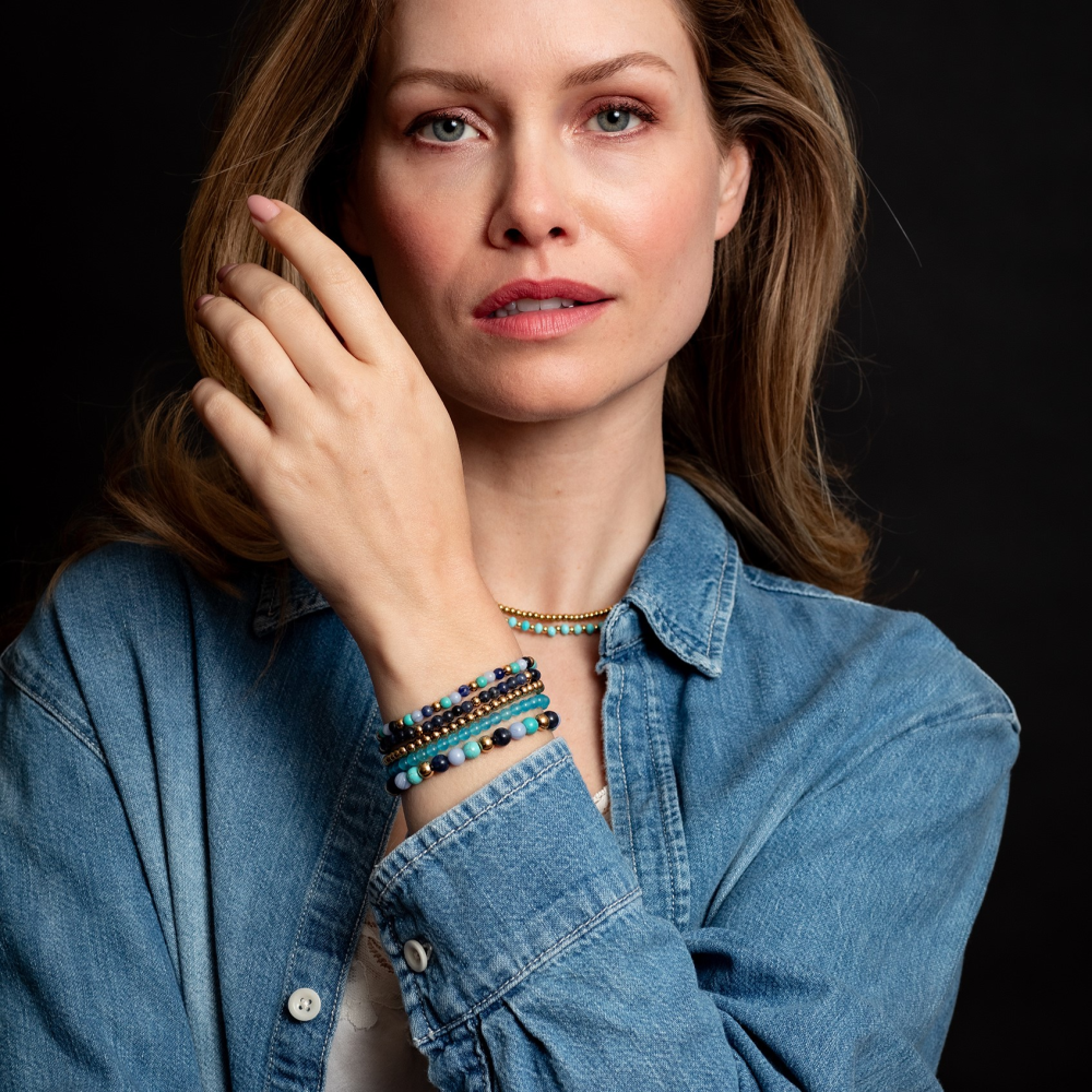 Een vrouw met lang lichtbruin haar in een denim shirt poseert tegen een donkere achtergrond. Ze draagt de Midnight Blue 4mm kralenarmband van Rebel & Rose en een 18k geelgouden geïoniseerde ketting.