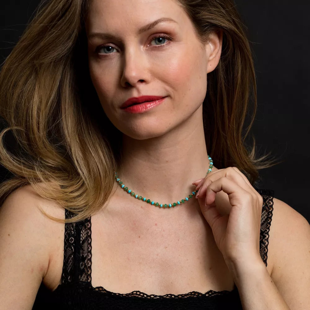 Een vrouw met lang, lichtbruin haar in een zwart kanten topje poseert tegen een donkere achtergrond. Ze houdt de Rebel & Rose Necklace Blue Green Adventure - 4mm vast en kijkt lichtjes opzij met een kalme uitdrukking.