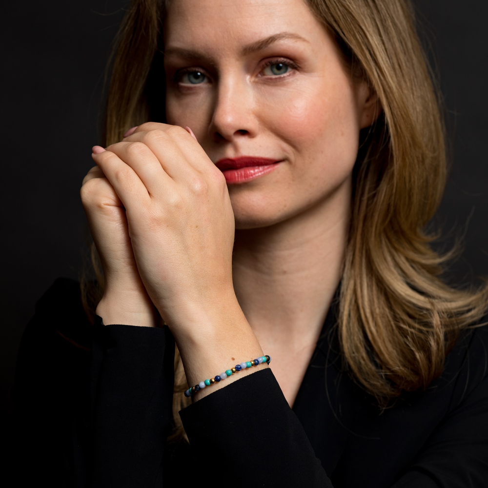 Een vrouw met lichtbruin haar in een zwart topje poseert tegen een donkere achtergrond, haar handen bij haar gezicht. Ze draagt de Rebel & Rose Winter Blues 18 kt geelgouden geïoniseerde 4mm kralen armband dames om haar pols.