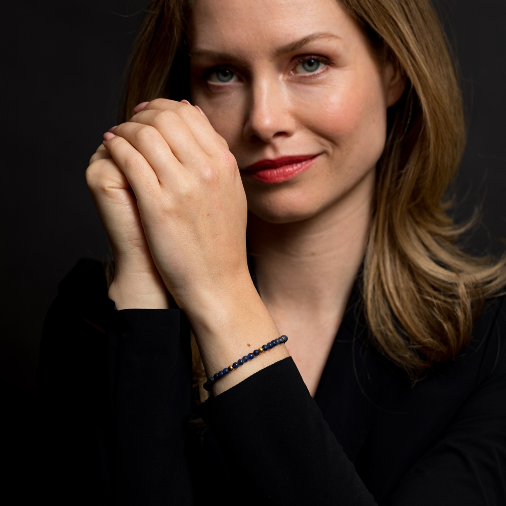 Een vrouw met lichtbruin haar en rode lippenstift draagt een zwart topje en de Rebel & Rose Midnight Blue 18 kt geelgouden geïoniseerde 4mm Kralen Armband Dames, poseert met haar handen bij haar gezicht tegen een donkere achtergrond.