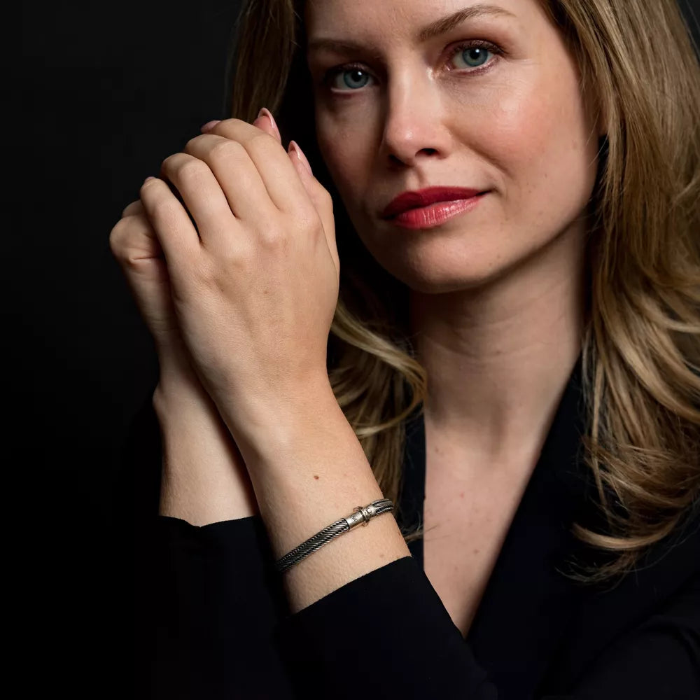 Een vrouw met lichtbruin haar en blauwe ogen poseert tegen een donkere achtergrond, draagt een zwart topje en een Rebel & Rose Demeter Small six - 17.11 gr. 925 armband, haar handen elegant om haar gezicht geklemd.