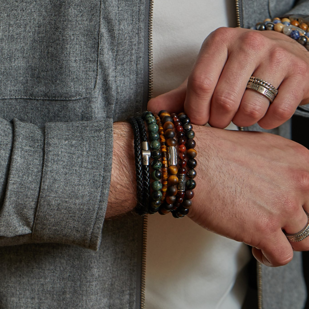 Een moderne man verstelt zijn Rebel & Rose Green Rocks - 8mm - Heren Kralen Armband en andere kralenarmbanden. Hij draagt een grijs jasje over een wit overhemd met ringen aan beide handen en straalt een verfijnde stijl uit.