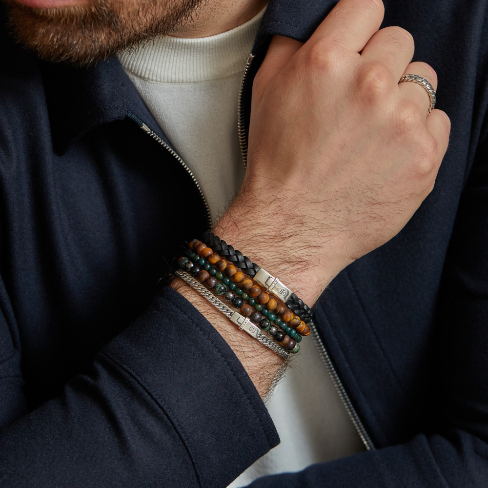 Een man met een donker jasje en wit overhemd benadrukt zijn pols met de Rebel & Rose Small Braided Raw Black - Leren Heren Armband, gecombineerd met andere kralen en metalen armbanden plus een zilveren ring voor een stijlvolle look.