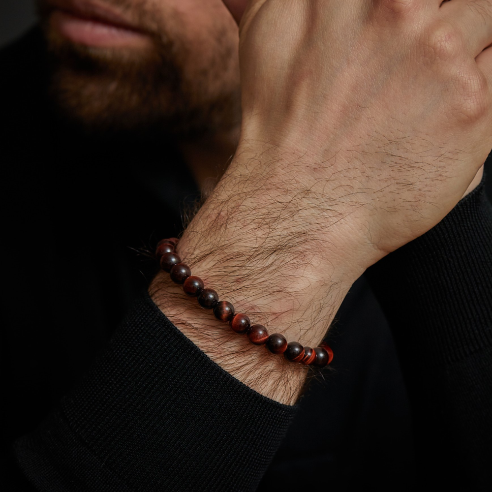 Een bebaard persoon in een zwart shirt met lange mouwen laat zijn kin op zijn hand rusten en toont een Rebel & Rose Tiger Red Vintage - 8mm - Heren Kralen Armband om zijn pols.