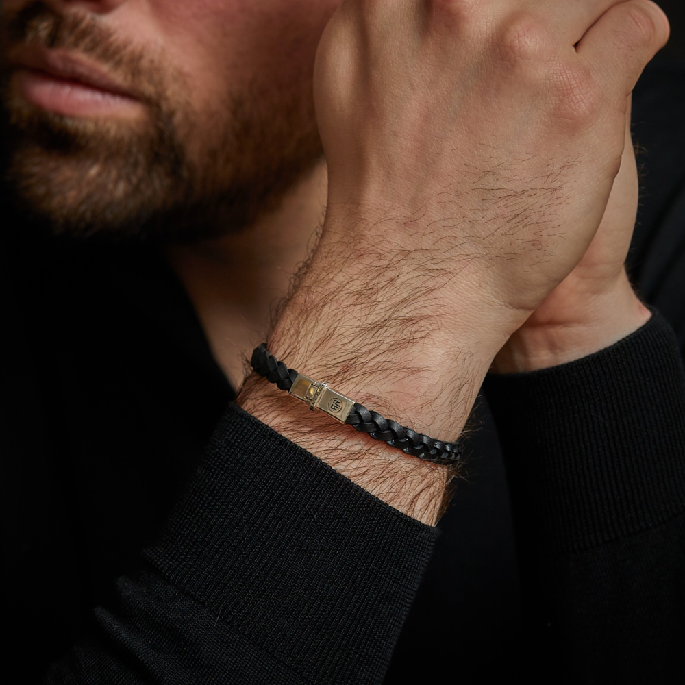 Een bebaarde man in een zwart shirt met lange mouwen laat zijn kin op zijn hand rusten en draagt de Rebel & Rose "Small Braided Raw Black - Leren Heren Armband," een zwarte leren armband met een zilveren sluiting.