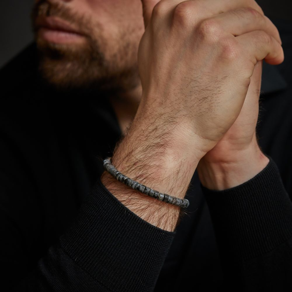 Een man in een donker shirt met lange mouwen omklemt zijn handen en toont de Rebel & Rose Slices - Grey Seduction 6mm heren kralen armband om zijn pols. Zijn gezicht is gedeeltelijk zichtbaar met een getrimde baard.