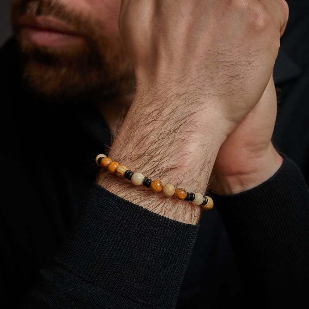 Een bebaarde man in een zwart shirt met lange mouwen houdt zijn handen bij zijn gezicht en draagt een Rebel & Rose kralen Woodstock - 8mm Kralen Heren Armband met gele, oranje, crème en zwarte kralen.