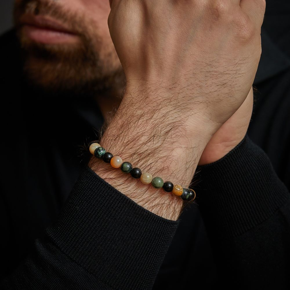 Een close-up van de handen van een bebaard persoon, die de Rebel & Rose Autumn Storm - 8mm Kralen Heren Armband met groene, zwarte en bruine stenen draagt, tegen een donkere achtergrond. De persoon is gekleed in een zwart shirt met lange mouwen.