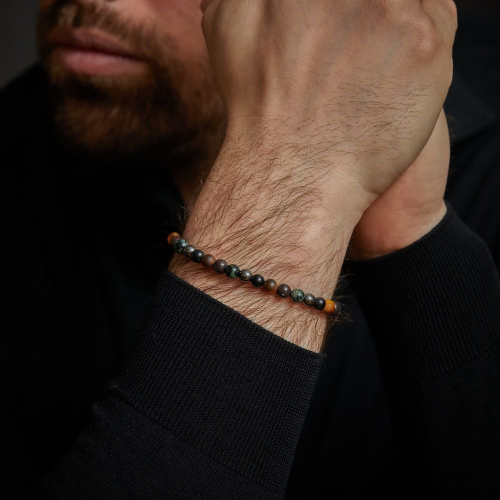 Een bebaarde man in donkere kleding kruist zijn armen en draagt een Rebel & Rose Stormy Weather Vintage - 6mm Heren Kralen Armband met donkere en bruine kralen om zijn pols.