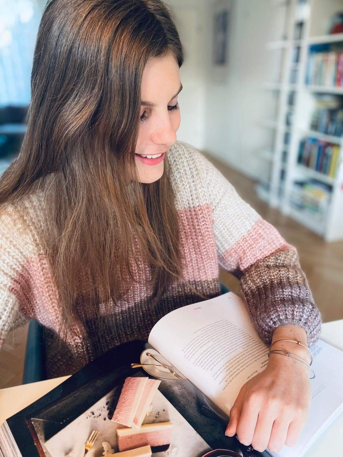 Een persoon met lang bruin haar in een gestreepte trui leest een boek aan een tafel met wafer cookies, een vork en de Thinking of You 🌸 armband basic van CLIC Creations. Op de achtergrond zijn wazige boekenplanken te zien.