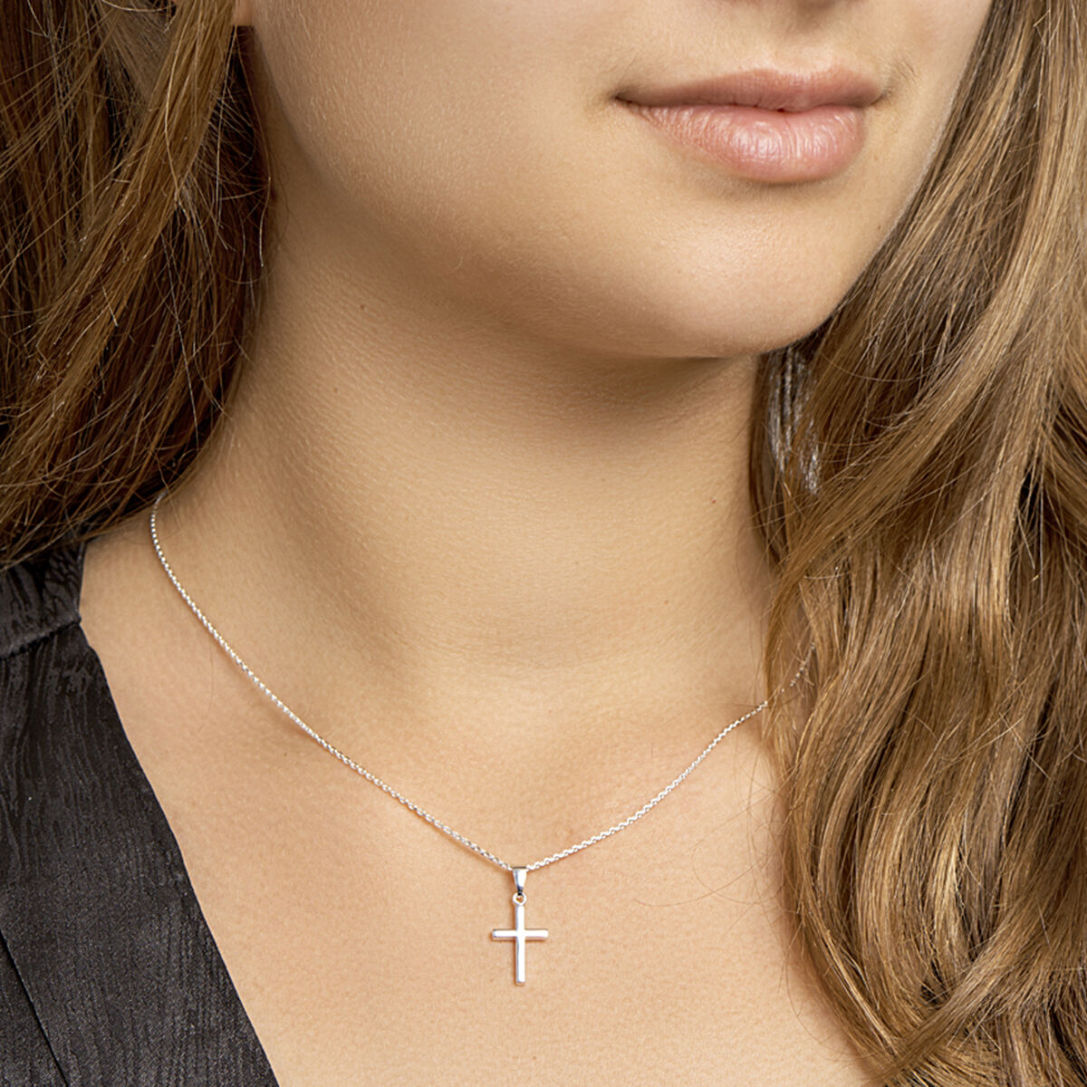 Een vrouw met lichtbruin haar draagt het Van der Kooij Collectie collier kruis 41 + 4 cm zilver wit, een fijne zilveren ketting met een klein kruisje. Gekleed in een top met donkere structuur zijn alleen haar lippen en kin gedeeltelijk zichtbaar voor een ingetogen look.